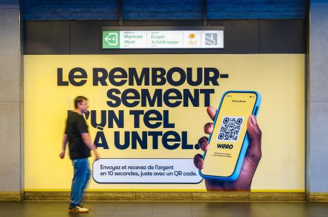 Een belevingswandaankleding (OOH) van JCDecaux Belgium in het Brusselse station De Brouckère zet de betaaldienst Wero in de kijker.  De felgele affiche toont de slogan « Le REMBOURSEMENT D'UN TEL À UNTEL. » en een smartphone met een QR-code, waar een voorbijganger in de gang van het station langsloopt.