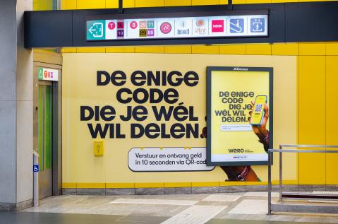 Een volledige station wrapping (OOH) van JCDecaux Belgium in het Brusselse metrostation De Brouckère zet de dienst Wero in de kijker.  De felgele wand toont de Nederlandse slogan « DE ENIGE CODE DIE JE WÉL WIL DELEN. » naast een verlicht paneel.