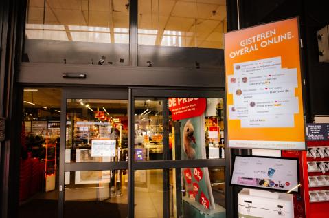 Un écran digital pDOOH de JCDecaux Belgium, est installé à l'entrée du magasin Carrefour à Etterbeek. Le visuel orange vif diffuse la campagne Aperol avec l'accroche « GISTEREN OVERAL ONLINE. » et des réactions de réseaux sociaux.