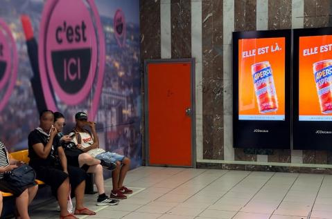 Un dispositif d'affichage digital programmatique (pDOOH) de l'univers MBT de JCDecaux Belgium, situé dans la station de métro « Louise » à Bruxelles, met en valeur une campagne publicitaire pour la marque Aperol.  Le double écran mural rétroéclairé diffuse un visuel orange vif présentant la canette Aperol Spritz surmontée du texte « ELLE EST LÀ. », devant une rangée de passagers assis sur des bancs le long d'un mur en marbre et d'une fresque murale.