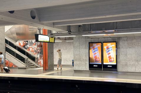 Un dispositif d'affichage digital programmatique (pDOOH) de JCDecaux Belgium, situé dans la station de métro « Porte de Namur » à Bruxelles, met en valeur une campagne publicitaire pour la marque Aperol.  Le double écran mural rétroéclairé diffuse un visuel orange vif présentant la canette Aperol Spritz surmontée du texte « FINI D’ATTENDRE. », tandis que des voyageurs attendent et circulent sur le quai le long des murs en marbre et des escaliers mécaniques.