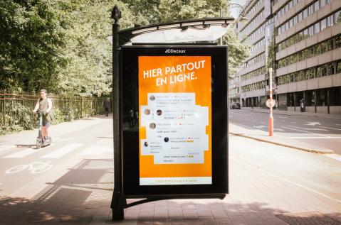 Un abribus digital de l'univers Street de JCDecaux Belgium, situé à Etterbeek (Bruxelles), diffuse une campagne publicitaire programmatique (pDOOH) pour la marque Aperol.  L'écran lumineux affiche un fond orange vif surmonté de l'accroche « HIER PARTOUT EN LIGNE. » et d'extraits de commentaires sociaux, à côté d'un usager en trottinette circulant le long d'une allée arborée.