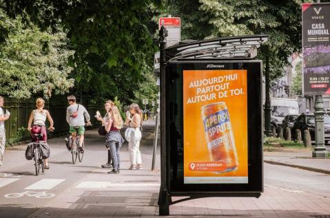 Un abribus digital (pDOOH) de JCDecaux Belgium, de l'univers Street à Etterbeek (Bruxelles), met en valeur une campagne publicitaire programmatique pour la marque Aperol. L'écran rétroéclairé diffuse un visuel orange vif affichant l'accroche « HIER PARTOUT EN LIGNE. » entourée d'extraits de commentaires sociaux, tandis qu'un usager en trottinette circule sur le trottoir le long d'une avenue arborée.