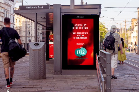 JCDecaux Belgium déploie une campagne programmatic DOOH sur un abribus à Anvers, dans l’univers Digital Street. Ce visuel rouge intense met en avant le slogan alertant sur la hausse des violences domestiques pendant les grands tournois, le numéro 1712 et une ambiance urbaine animée par le passage de piétons.