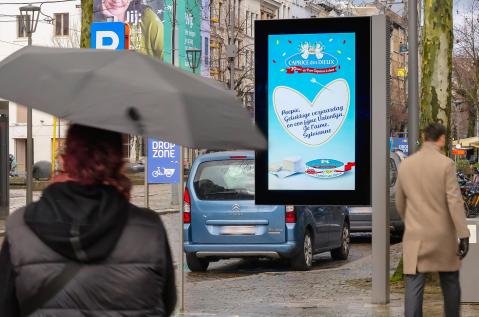 Een digitaal stedelijk reclamescherm met een valentijnsboodschap van "Caprice des Dieux". Binnen een wit hart is een persoonlijke liefdes- en verjaardagsboodschap te lezen: "Poepie, Gelukkige verjaardag en een fijne Valentijn Je t'aime. Sylvianne". Op de achtergrond en rondom het scherm fietst een fietser in een oranje shirt langs een tramhalte waar een tram stilstaat, staat een blauwe auto vlakbij geparkeerd en maken gevels van gebouwen het stadsdecor compleet onder winterse bomen.