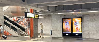 Een programmatic DOOH-display (pDOOH) van JCDecaux Belgium in het Brusselse metrostation 'Naamsepoort' zet een reclamecampagne voor het merk Aperol in de kijker.  Het dubbele verlichte wandscherm toont een feloranje visual van het Aperol Spritz-blikje met daarboven de slogan « FINI D’ATTENDRE. », terwijl reizigers wachten en zich verplaatsen op het perron langs de marmeren wanden en de roltrappen.