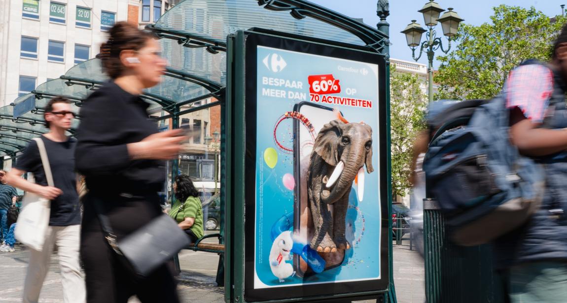 Photographie urbaine montrant un abribus avec un panneau publicitaire lumineux Carrefour. Des passants flous marchent au premier plan. Sur l'affiche, sur fond bleu ciel, un grand smartphone montre un éléphant d'attraction et un Lapin Crétin semblant surgir de l'écran, entourés de ballons et d'un tracé de montagnes russes. Le slogan en néerlandais indique : « BESPAAR TOT 60% OP MEER DAN 70 ACTIVITEITEN » (Économisez jusqu'à 60 % sur plus de 70 activités).