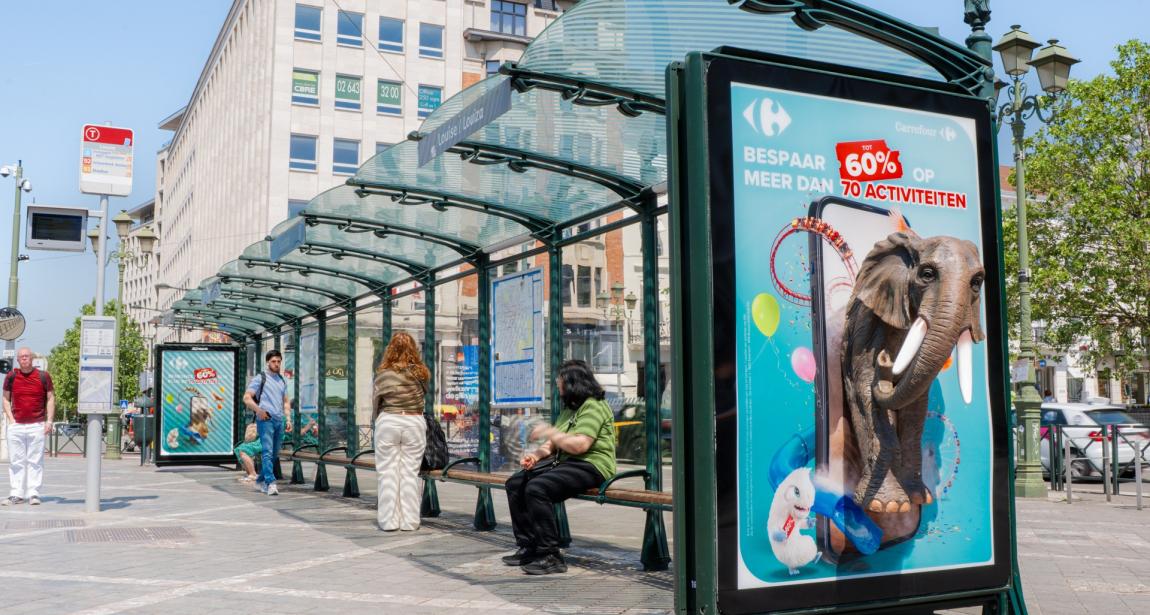Vue d’ensemble en perspective d’une grande station d’abribus vitrée sous un ciel bleu ensoleillé. Au premier plan, un panneau d'affichage lumineux présente une campagne publicitaire Carrefour en néerlandais (« BESPAAR TOT 60% OP MEER DAN 70 ACTIVITEITEN »). L'affiche intègre un effet 3D spectaculaire avec une tête d'éléphant grandeur nature qui ressort physiquement de la paroi, accompagnée d'un Lapin Crétin et d'un smartphone sur fond bleu ciel. Sous l’abri, des usagers attendent assis ou debout sur le trot