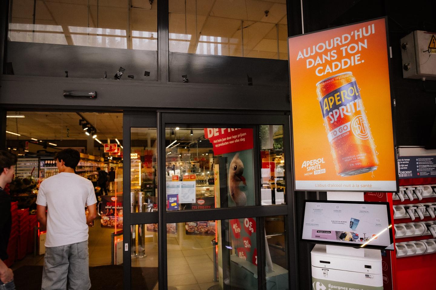 Un écran digital programmatique (pDOOH) de l'univers Retail de JCDecaux Belgium, situé à l'entrée d'un supermarché Carrefour à Etterbeek (Bruxelles), met en valeur une campagne publicitaire pour la marque Aperol.  Le panneau vertical rétroéclairé diffuse un visuel orange vif mettant en avant une canette Aperol Spritz accompagnée du slogan « AUJOURD'HUI DANS TON CADDIE. », placé à côté d'une borne interactive et de clients franchissant les portes automatiques.