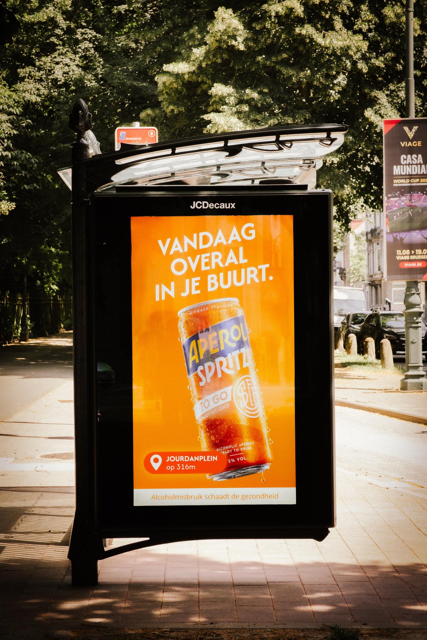 Un abribus digital (pDOOH) de l'univers Street de JCDecaux Belgium, situé à Bruxelles, met en valeur une campagne publicitaire programmatique pour la marque Aperol.  L'écran rétroéclairé diffuse un visuel orange vif mettant en avant une canette Aperol Spritz avec le texte en néerlandais « VANDAAG OVERAL IN JE BUURT. » et une géolocalisation « JOURDANPLEIN op 316m », sur un trottoir pavé bordé d'arbres.