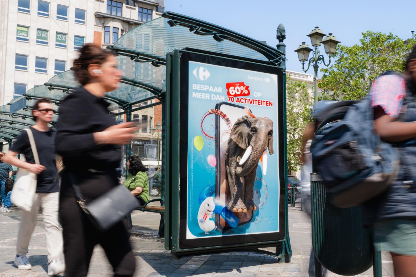 Photographie urbaine montrant un abribus avec un panneau publicitaire lumineux Carrefour. Des passants flous marchent au premier plan. Sur l'affiche, sur fond bleu ciel, un grand smartphone montre un éléphant d'attraction et un Lapin Crétin semblant surgir de l'écran, entourés de ballons et d'un tracé de montagnes russes. Le slogan en néerlandais indique : « BESPAAR TOT 60% OP MEER DAN 70 ACTIVITEITEN » (Économisez jusqu'à 60 % sur plus de 70 activités).