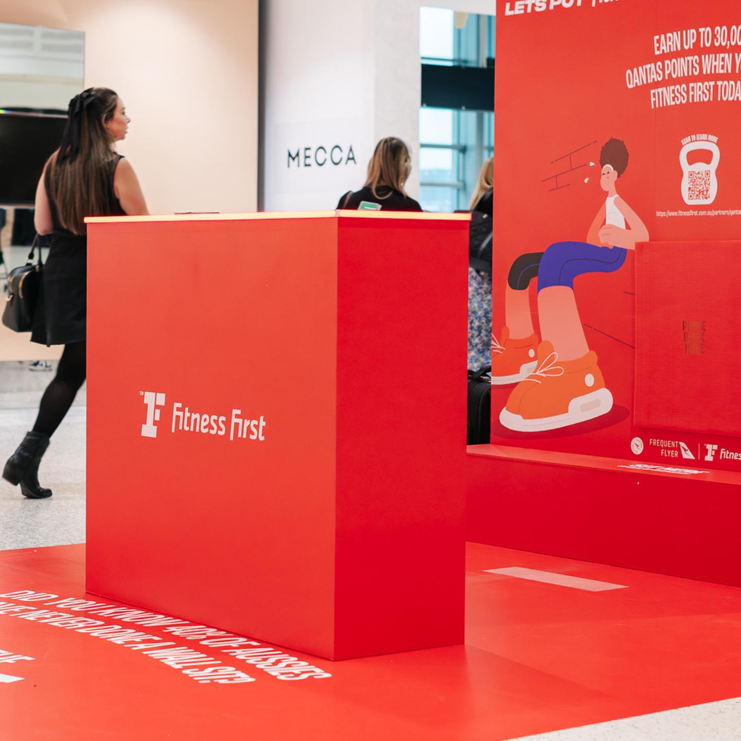 Wide shot of the Fitness First campaign space in the airport concourse, showing the branded red structure integrated into the JCDecaux environment and engaging passing travellers.