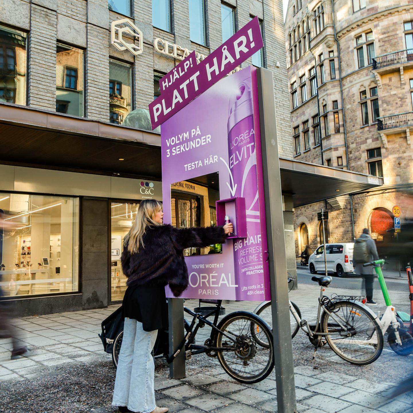 Woman standing in front of a JCDecaux digital mirror, trying L'Oréal Elvital Collagen Lifter spray and seeing the volumising effect on her hair in real time.
