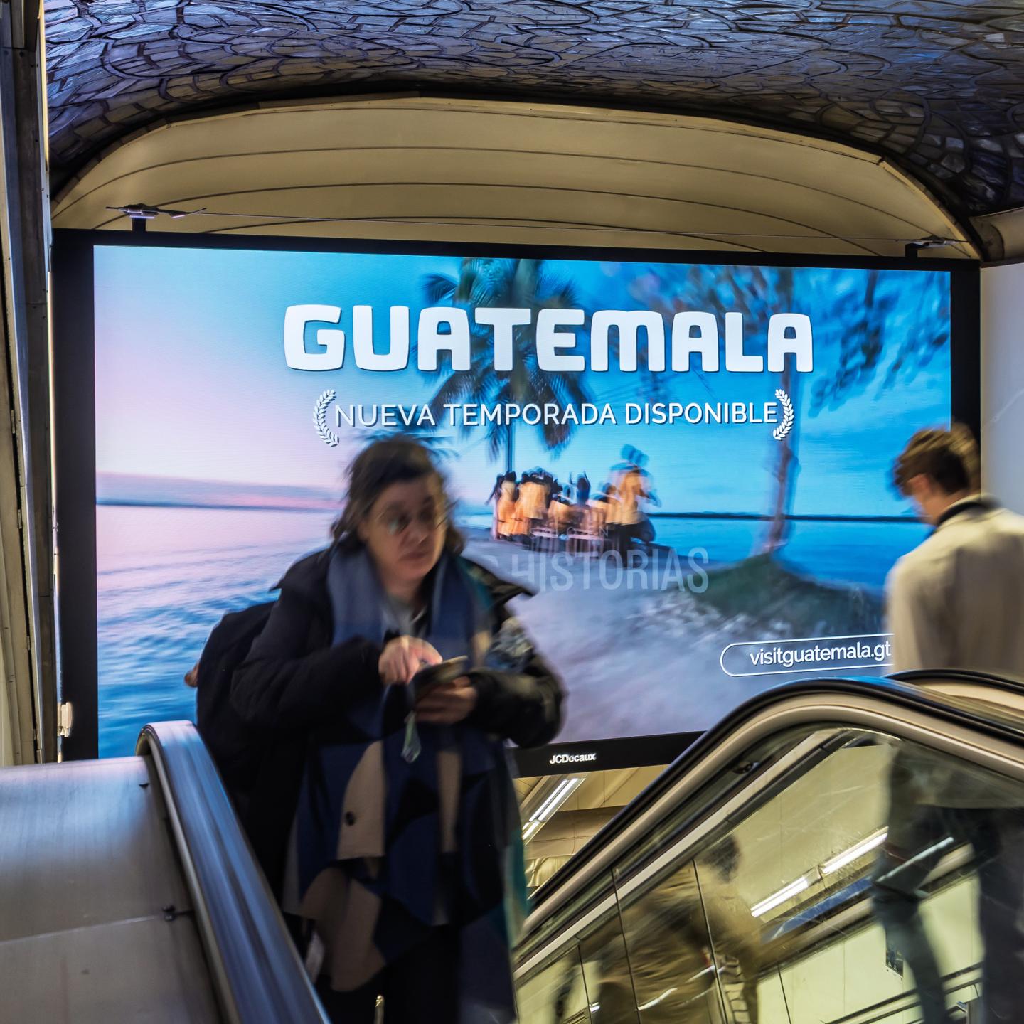 Travellers walking through a Madrid metro corridor transformed by JCDecaux Spain into a colourful gateway to Guatemala with lush landscape visuals.