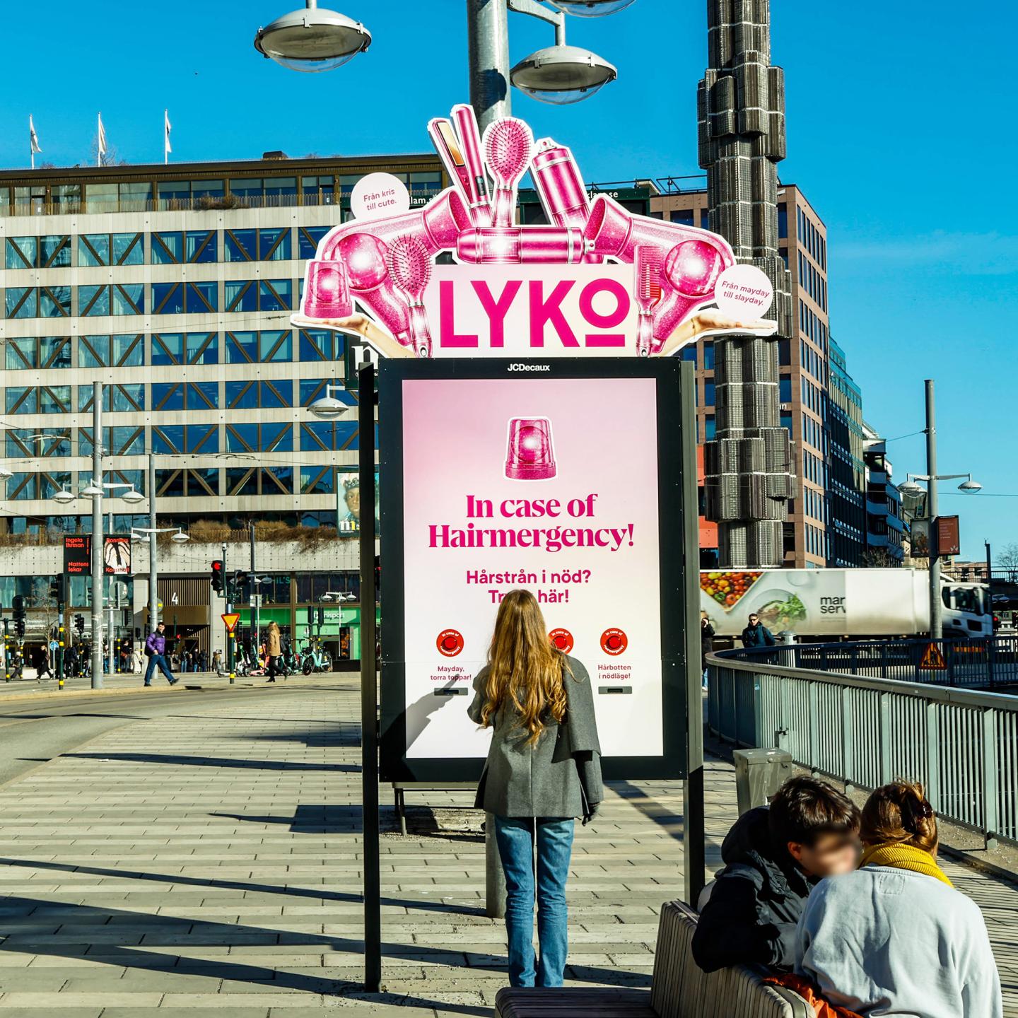 Woman standing in front of a JCDecaux digital bus shelter displaying a pink Lyko “In case of Hairmergency” advert in central Stockholm