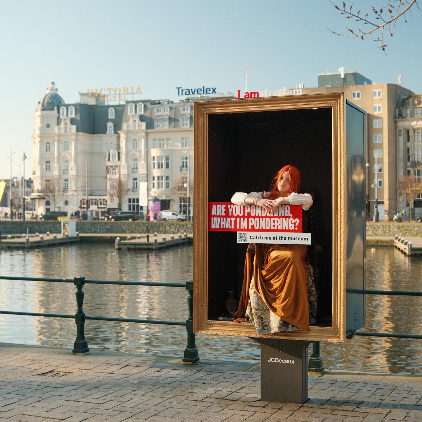 A performer dressed as a painting character standing inside a street furniture display near Amsterdam Central Station.