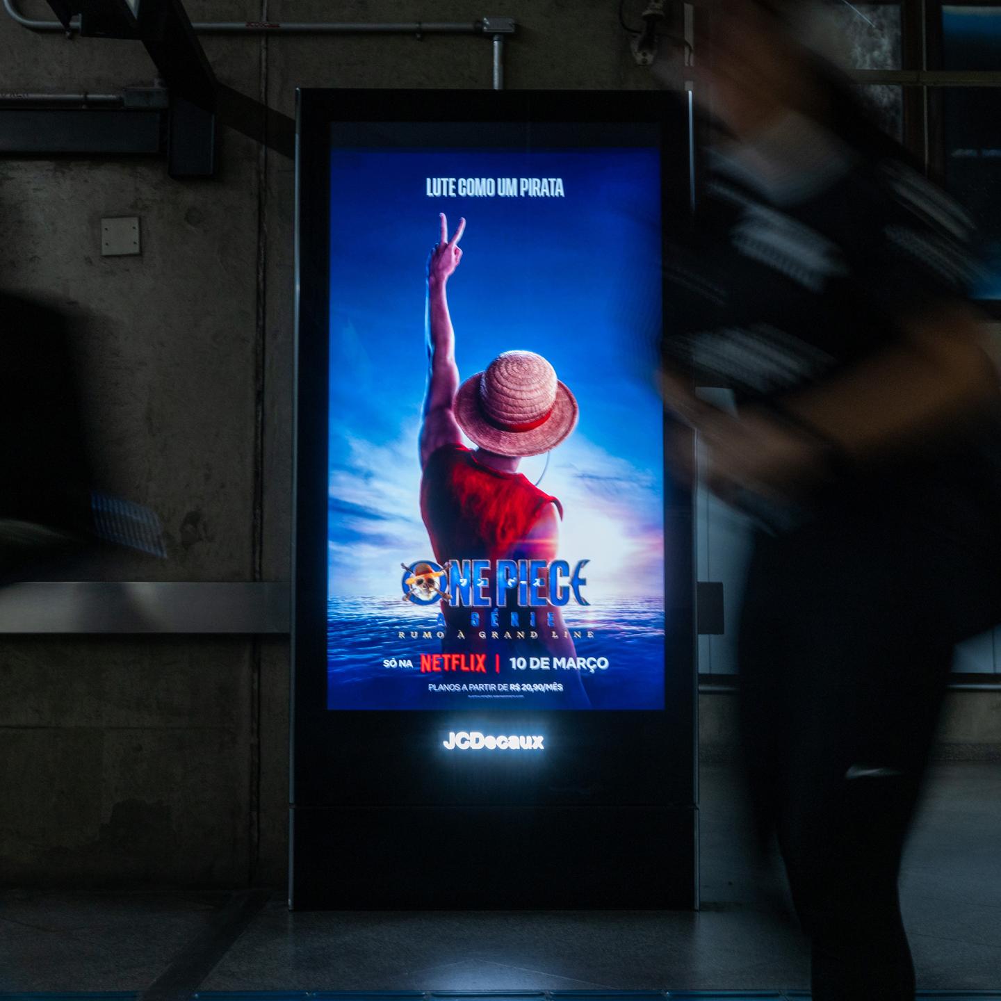 Commuters walking through a São Paulo metro station featuring a dramatic One Piece Season 2 display with Luffy’s arms extending across the space.