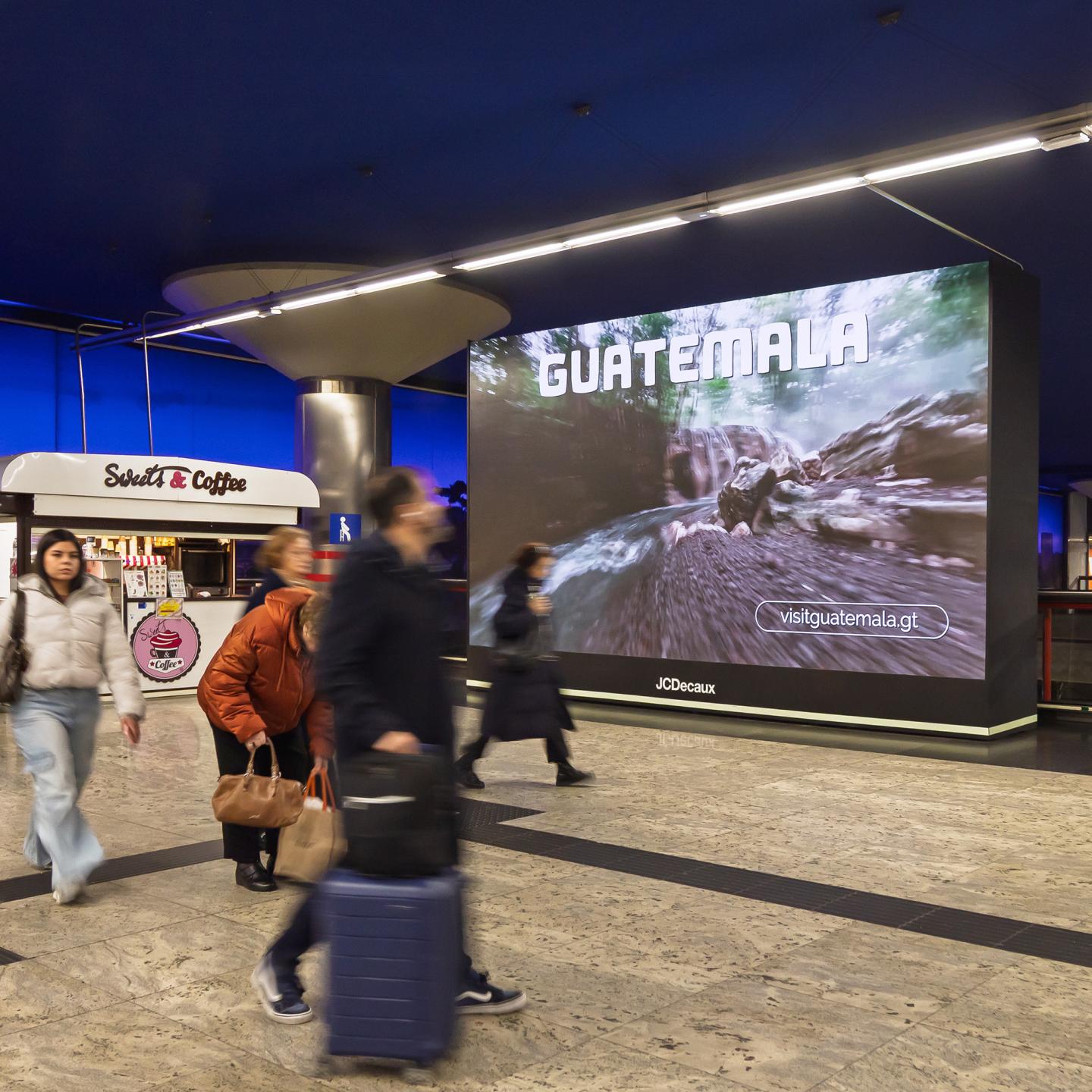 Passengers passing bright Visit Guatemala posters along a metro walkway, invited to explore Guatemala’s nature and heritage.