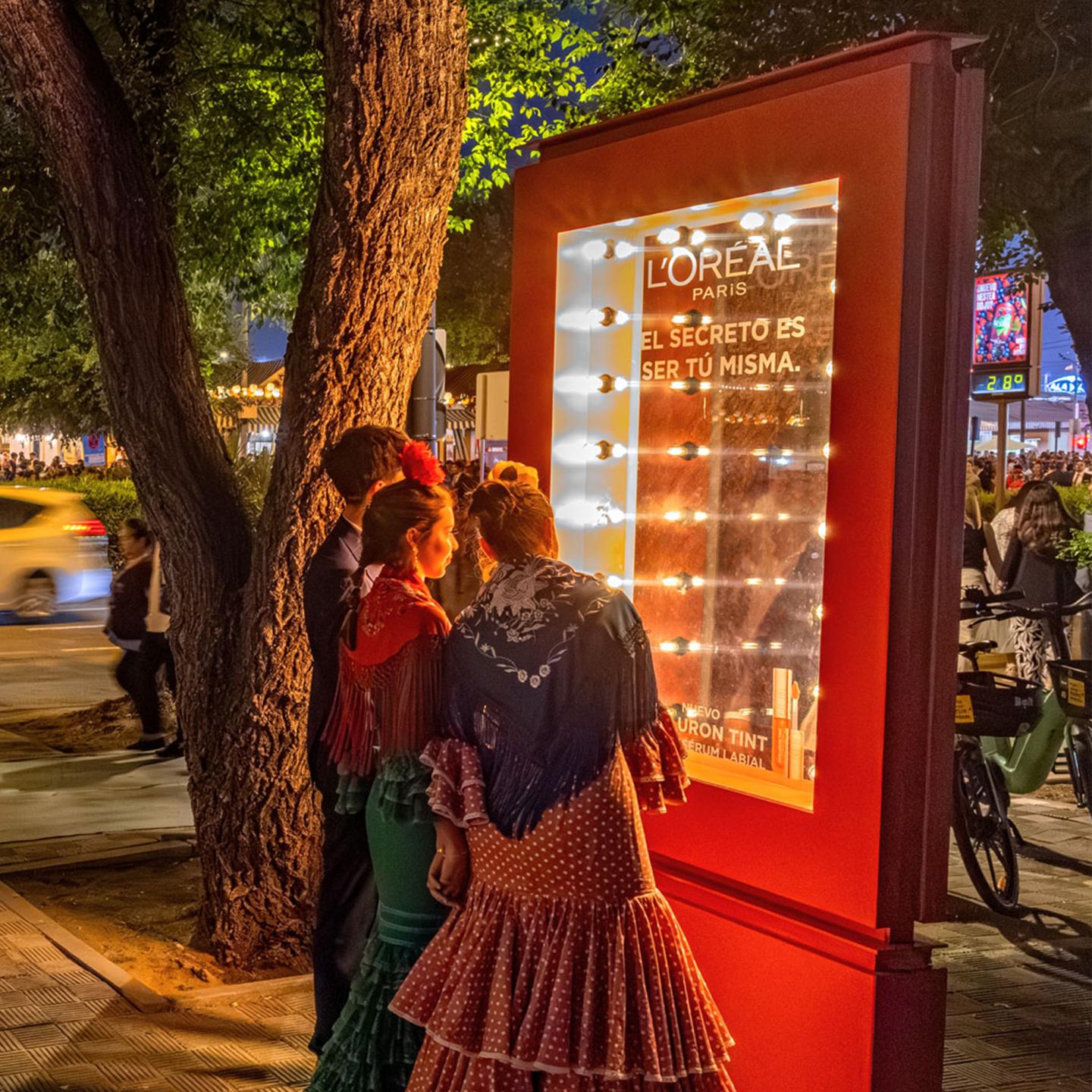Two women in traditional Sevillana dresses looking into an illuminated L’Oréal Paris vanity mirror installed by JCDecaux in Seville.