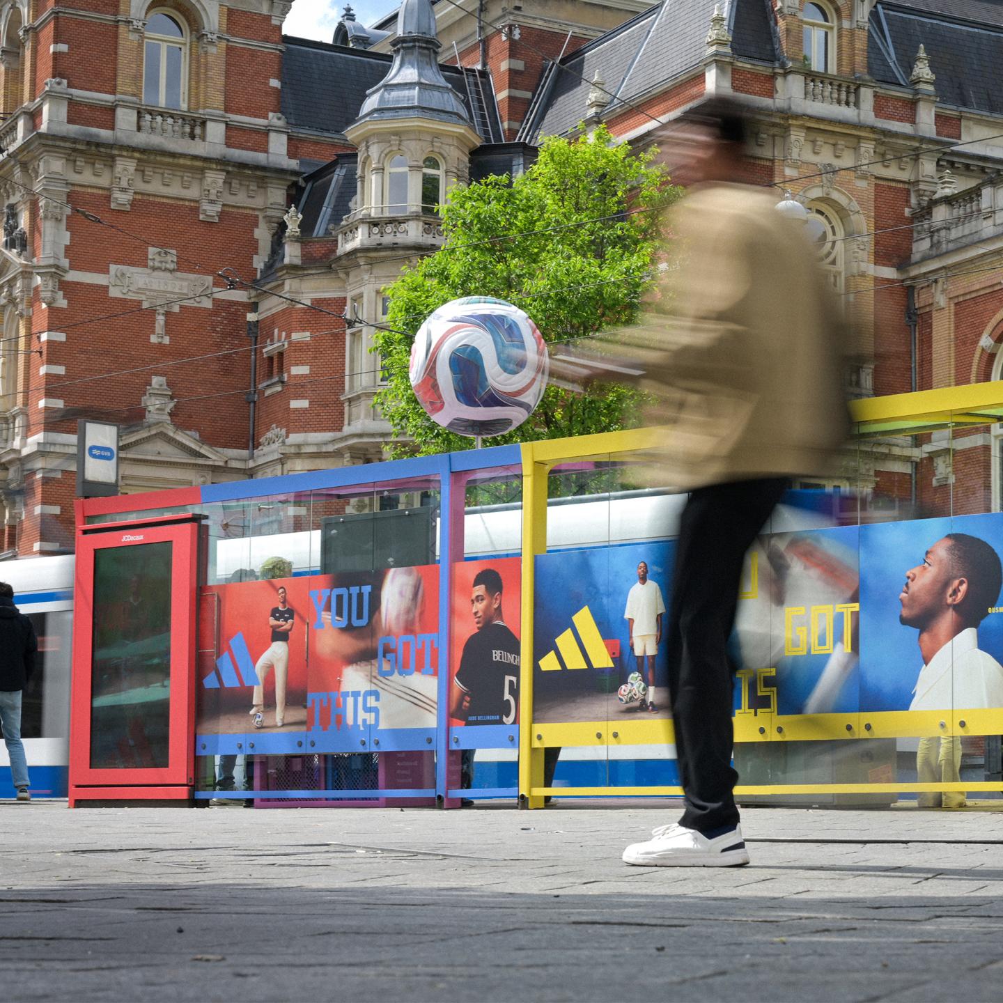 Pedestrian walking past an adidas World Cup special build on Amsterdam street furniture, featuring large images of international football stars.