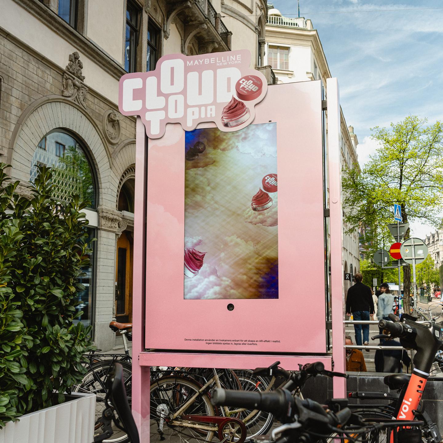 Interactive JCDecaux street furniture displaying the Maybelline Cloudtopia campaign, showing an AR beauty experience on a pink digital screen in a busy Stockholm shopping street.