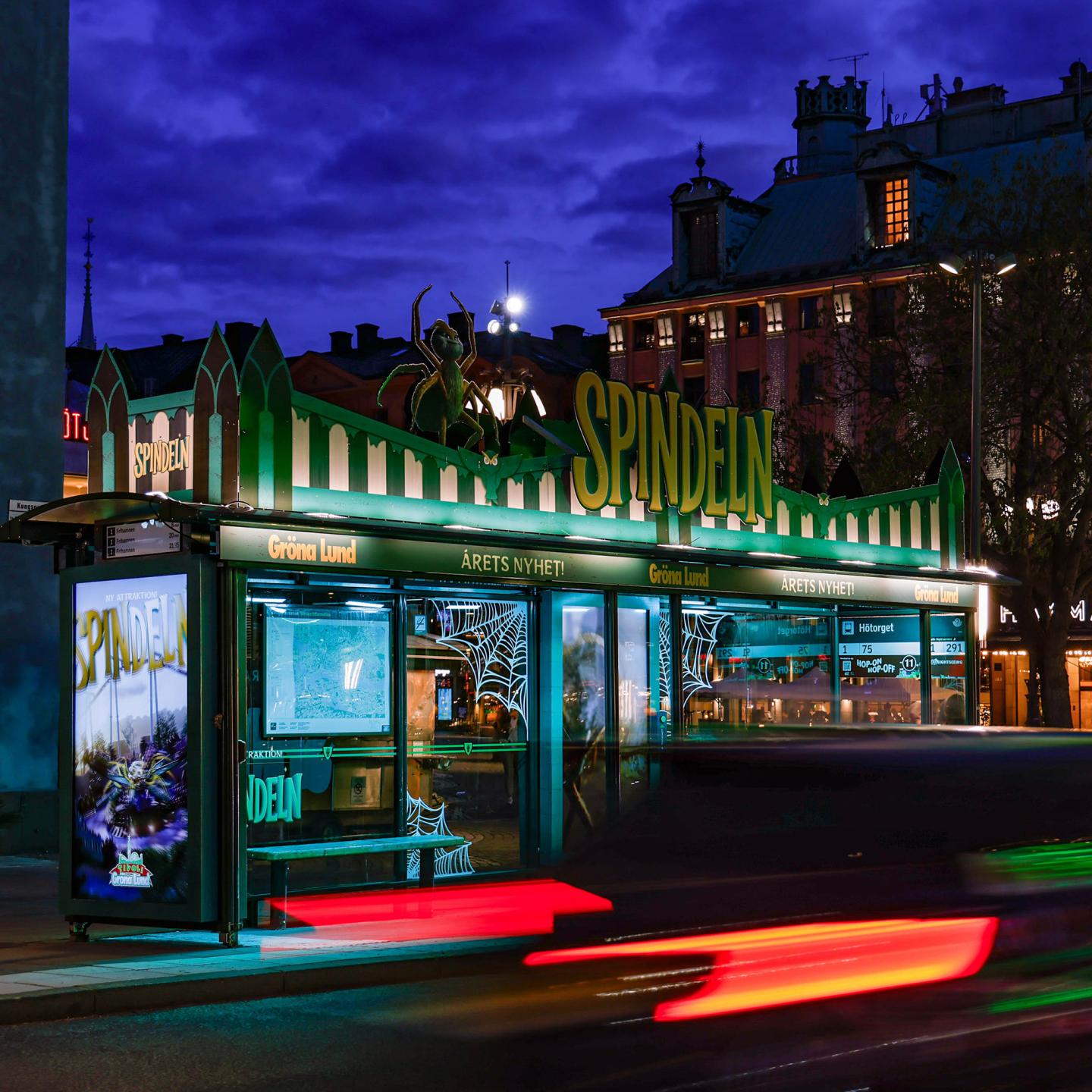 Wide shot of a street in Stockholm featuring a bus shelter fully branded in dark colours with a monumental spider installation.