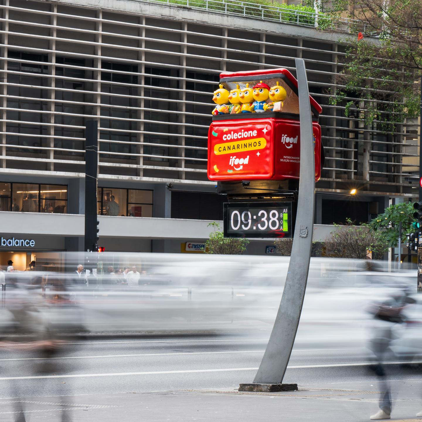 JCDecaux digital clock on Paulista Avenue showing the iFood Canaries plush characters above the screen, surrounded by pedestrians and office buildings.