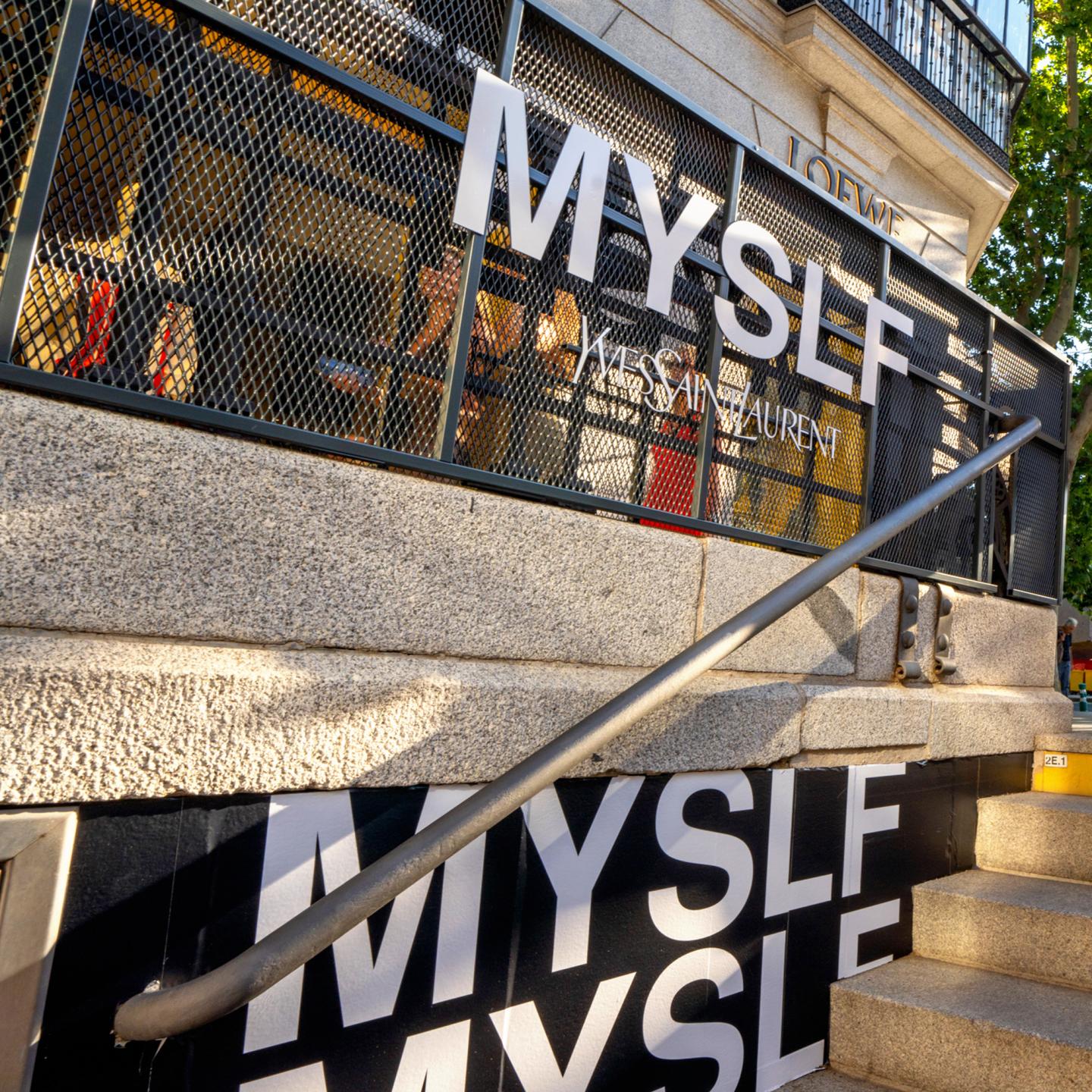 Wide shot of the Serrano metro access branded by JCDecaux, showing repeated MYSLF logos and large perfume imagery along the railings and walls.