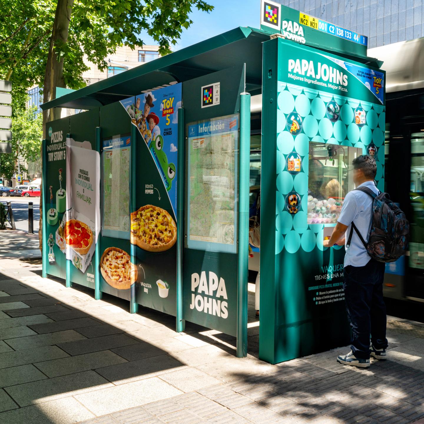 Close-up of a JCDecaux bus shelter in Madrid equipped with an interactive claw machine, offering Toy Story 5 merchandise and Papa Johns pizza prizes to passers-by.