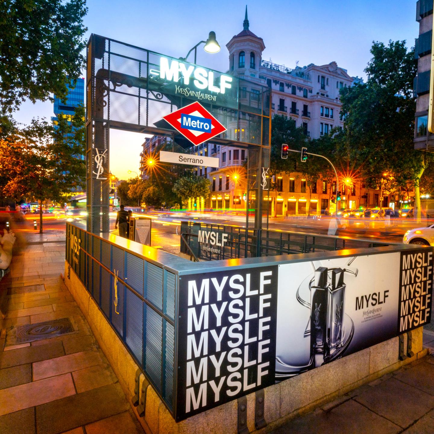 Night view of Serrano metro entrance in Madrid, illuminated and fully wrapped in MYSLF visuals by JCDecaux, with city lights and traffic in the background.