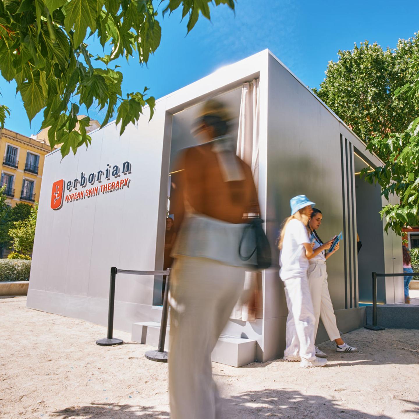 Street view of the JCDecaux and Erborian glow skincare pop-up in central Madrid, with pedestrians walking by the modern metallic structure under the trees.