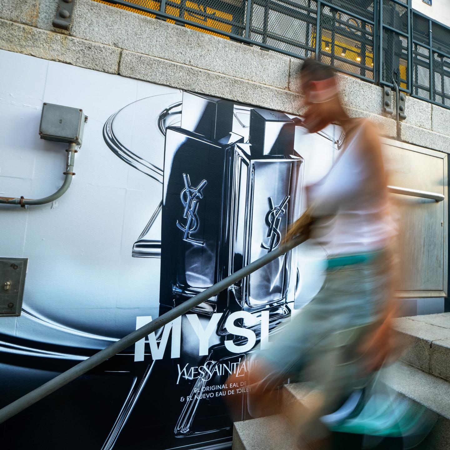 Large JCDecaux wall display featuring the MYSLF fragrance bottle by Yves Saint Laurent, with a blurred pedestrian walking past on the station steps.