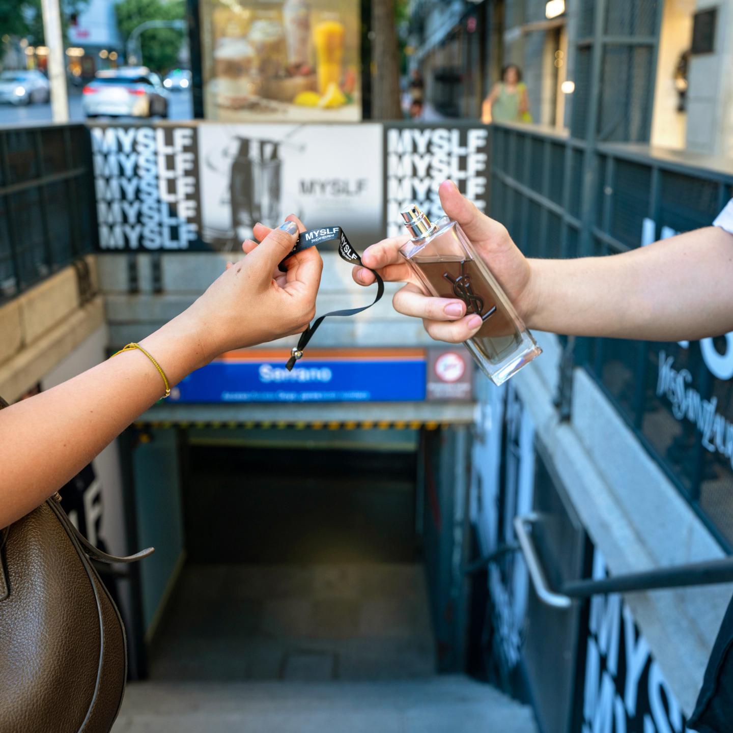 Close‑up of two hands exchanging a bracelet infused with MYSLF fragrance in front of the JCDecaux‑branded Serrano metro entrance.