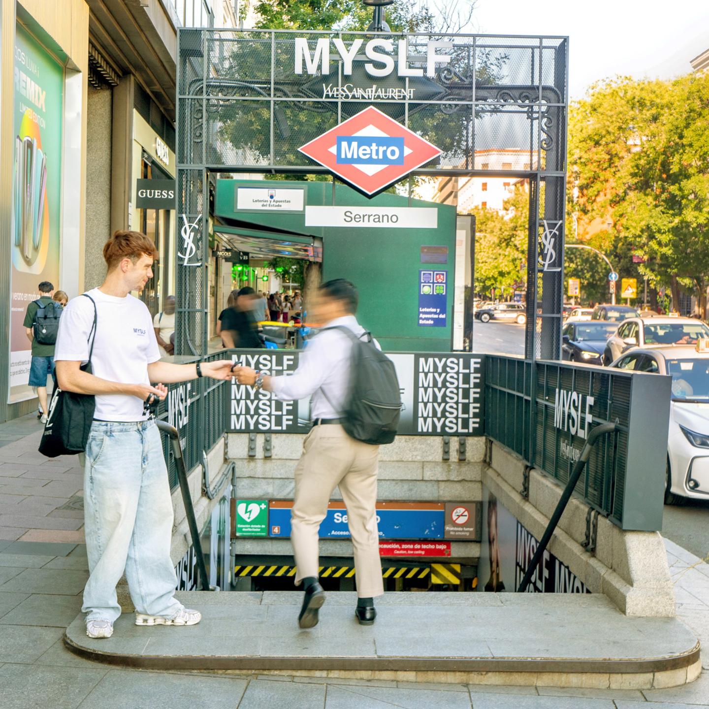 Commuters entering the JCDecaux‑branded Serrano metro station, with MYSLF signage and black‑and‑white graphics surrounding the staircase and entrance.