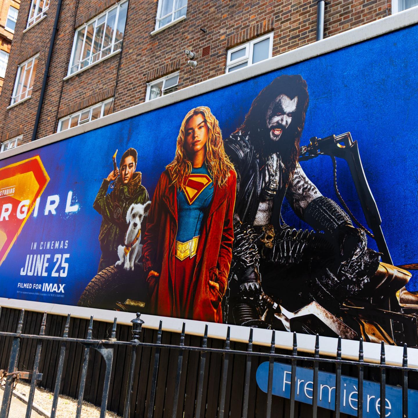 Close‑up of a large Supergirl out‑of‑home billboard featuring a tilted 3D logo and vibrant colours on a London street.