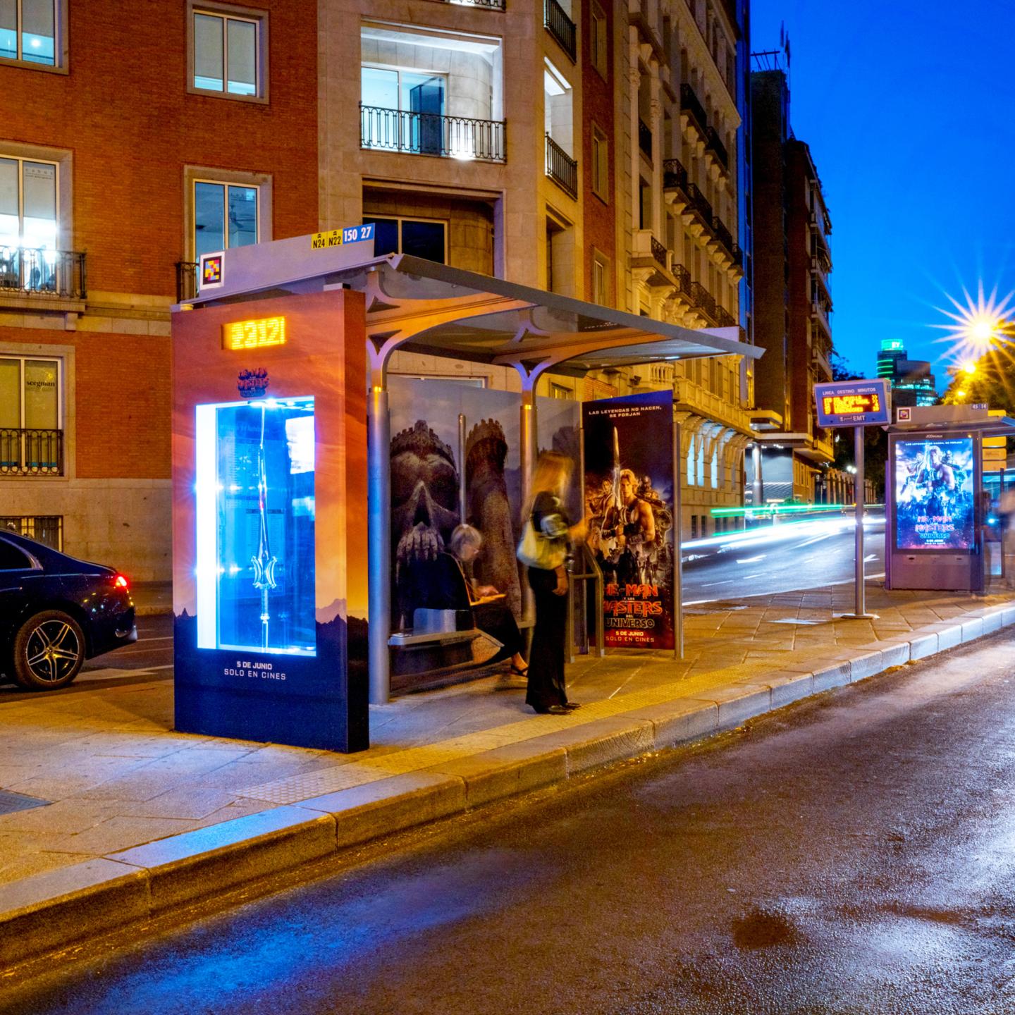 JCDecaux bus shelter fully wrapped in Masters of the Universe artwork, showing film characters and dramatic skyline in a busy Madrid street.