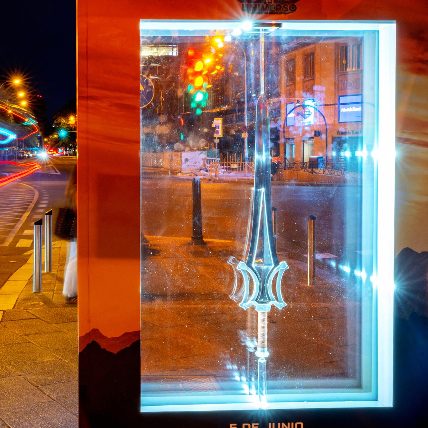 Close-up of the backlit Power Sword replica displayed inside JCDecaux street furniture on a Madrid avenue.