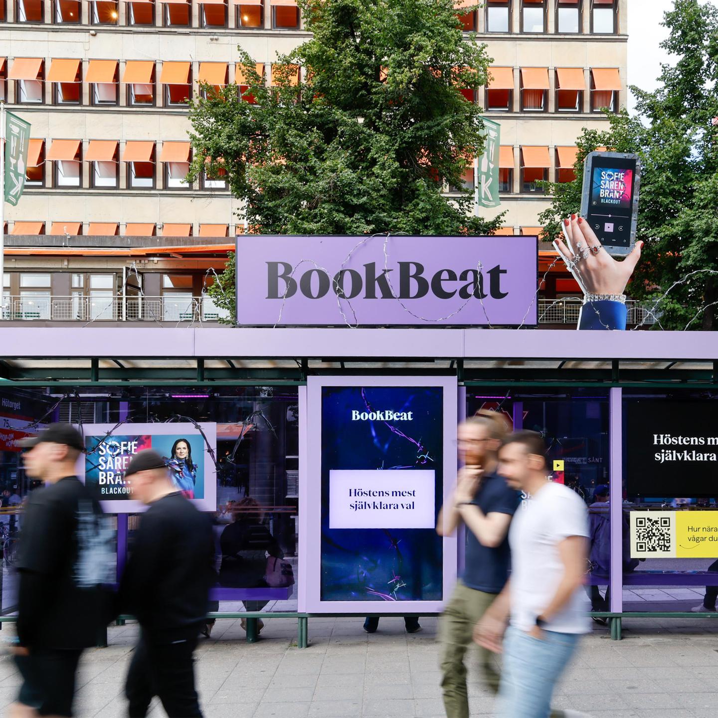 Street view of a BookBeat-branded on JCDecaux bus shelter in central Stockholm, featuring bright neon reflections and outdoor advertising for audiobooks.
