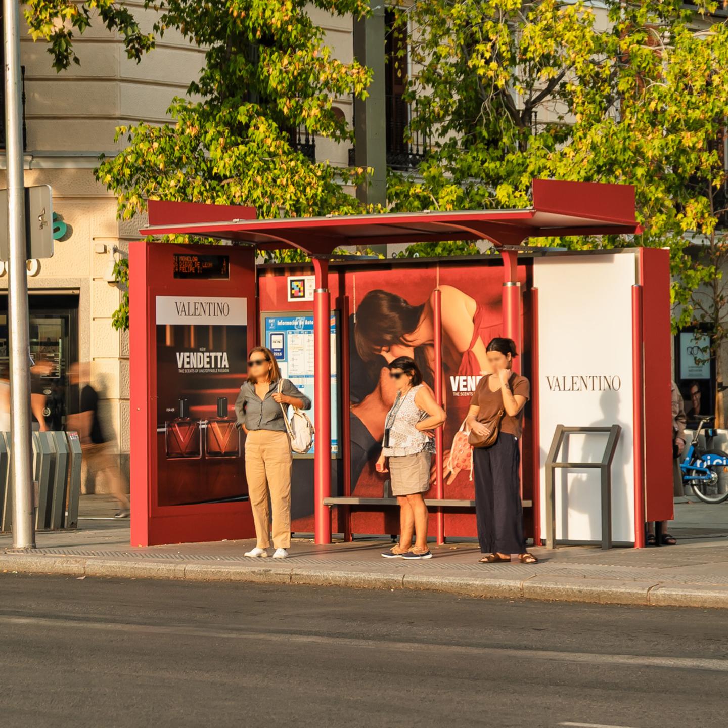 Three women waiting at a JCDecaux Spain bus shelter in Madrid, fully wrapped in Valentino Vendetta branding with perfume and couple visuals.