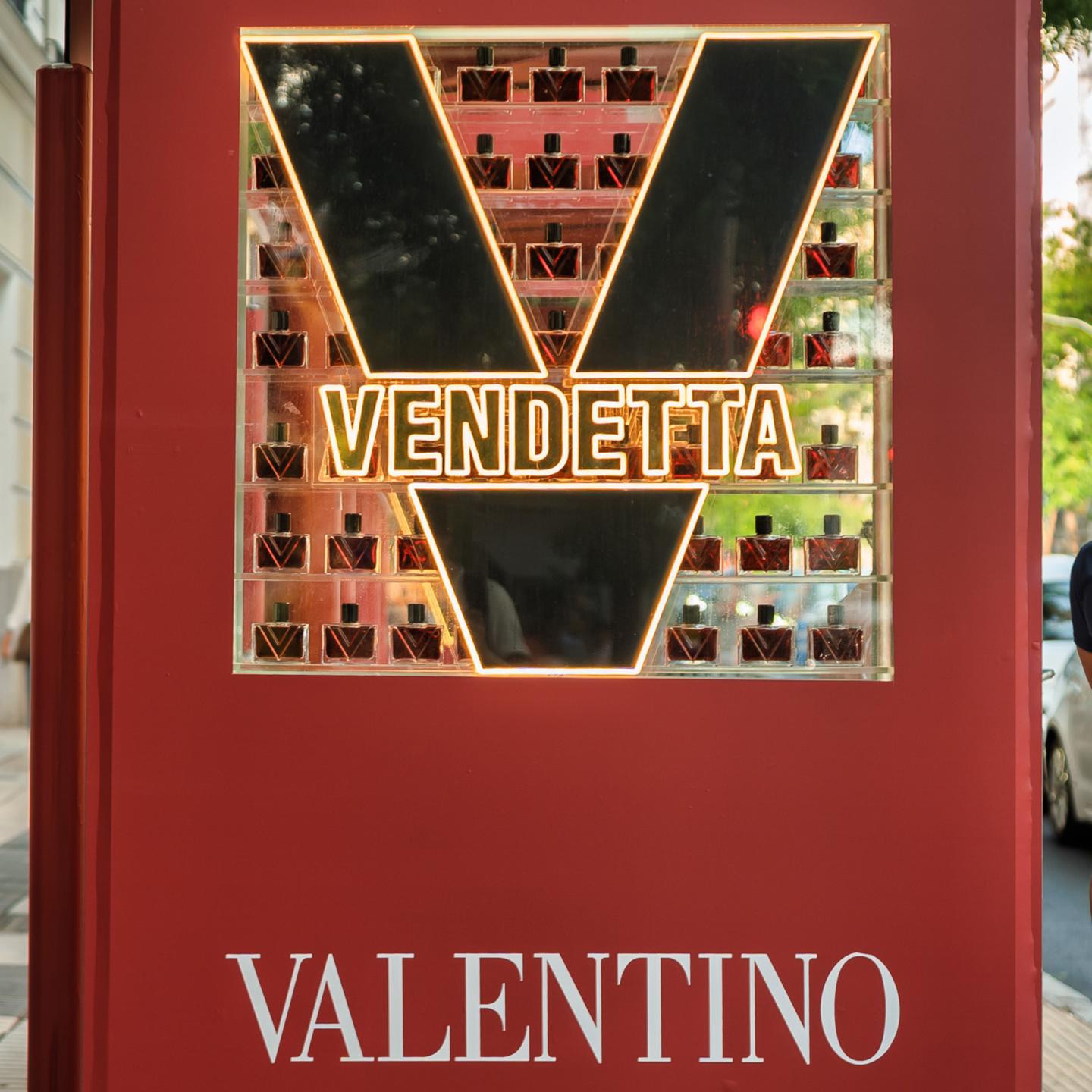 Close-up of a red JCDecaux Spain street furniture unit in Madrid featuring a glowing V-shaped Valentino Vendetta logo filled with rows of perfume bottles.
