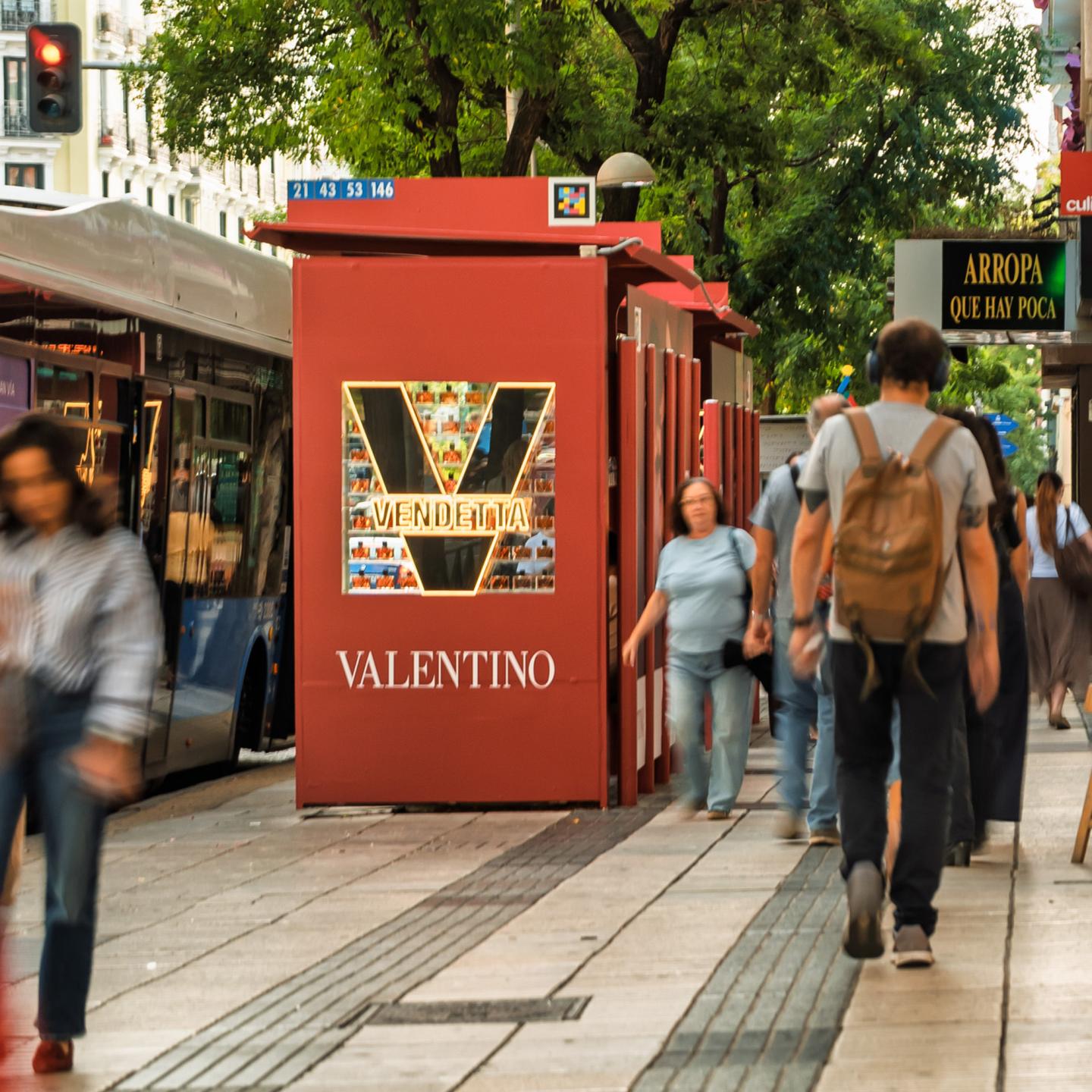 Valentino Vendetta campaign displayed on a red JCDecaux Spain bus shelter along a crowded Madrid avenue, surrounded by passing pedestrians and buses.