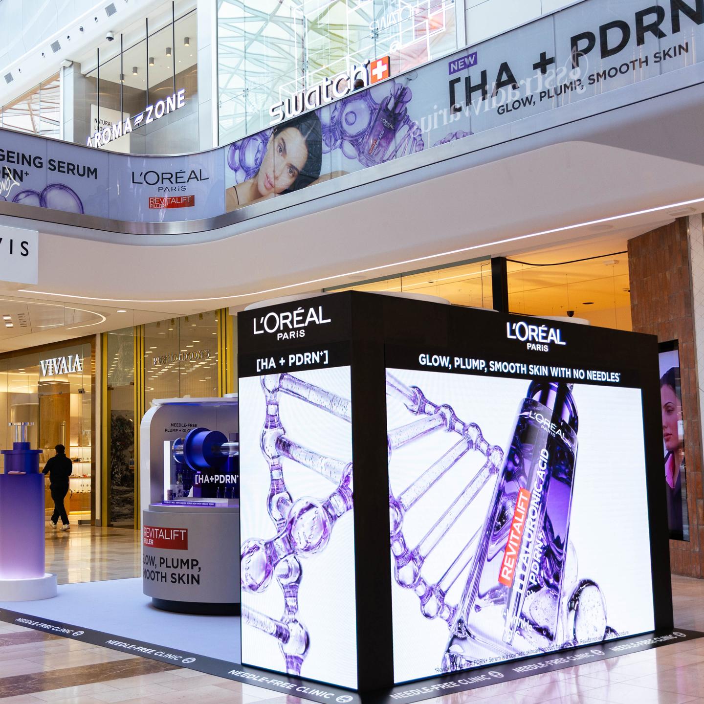 Bright digital cube showing L’Oréal Revitalift [HA + PDRN] serum visuals in the mall walkway at Westfield London, surrounded by retail stores and overhead campaign banners.