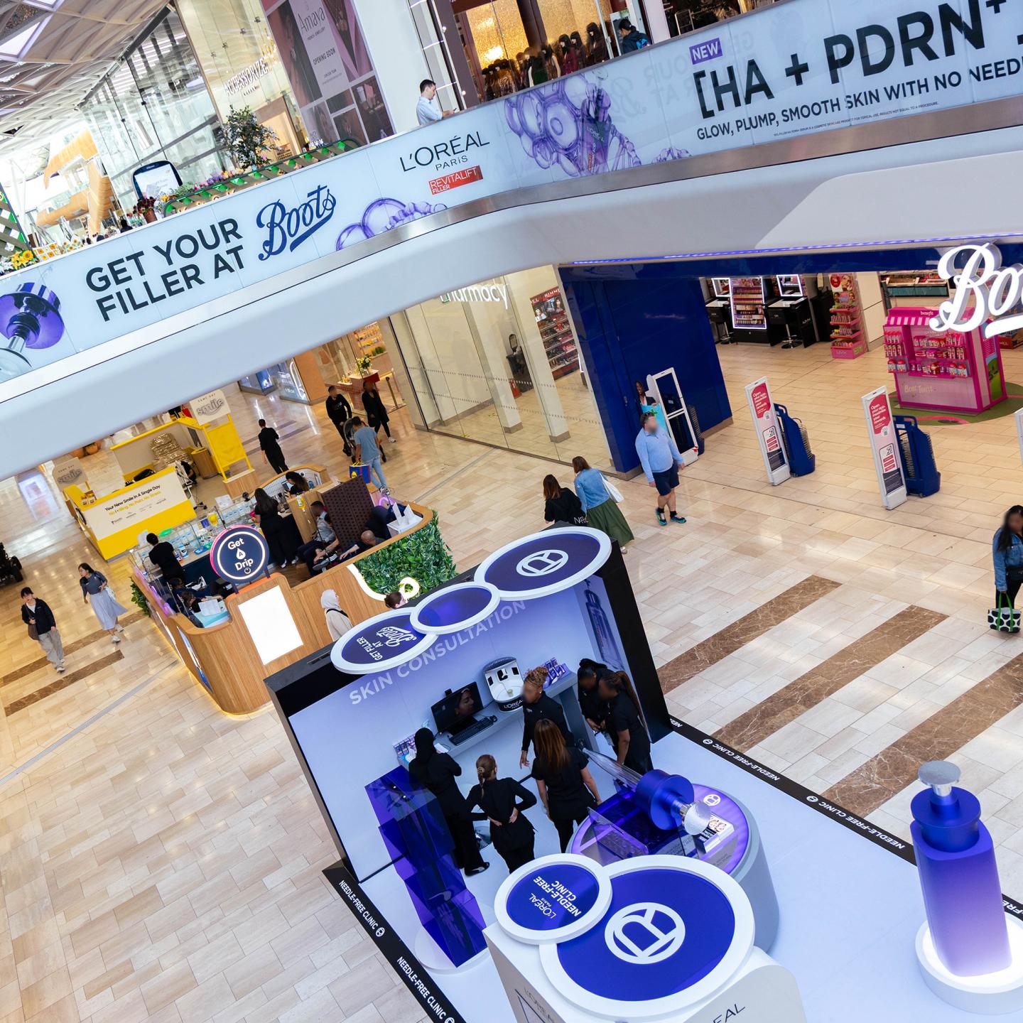 Overhead view of a L’Oréal Revitalift skin consultation pop-up and promotional banners inside Westfield London, with shoppers walking past Boots and other retail stores in the mall.