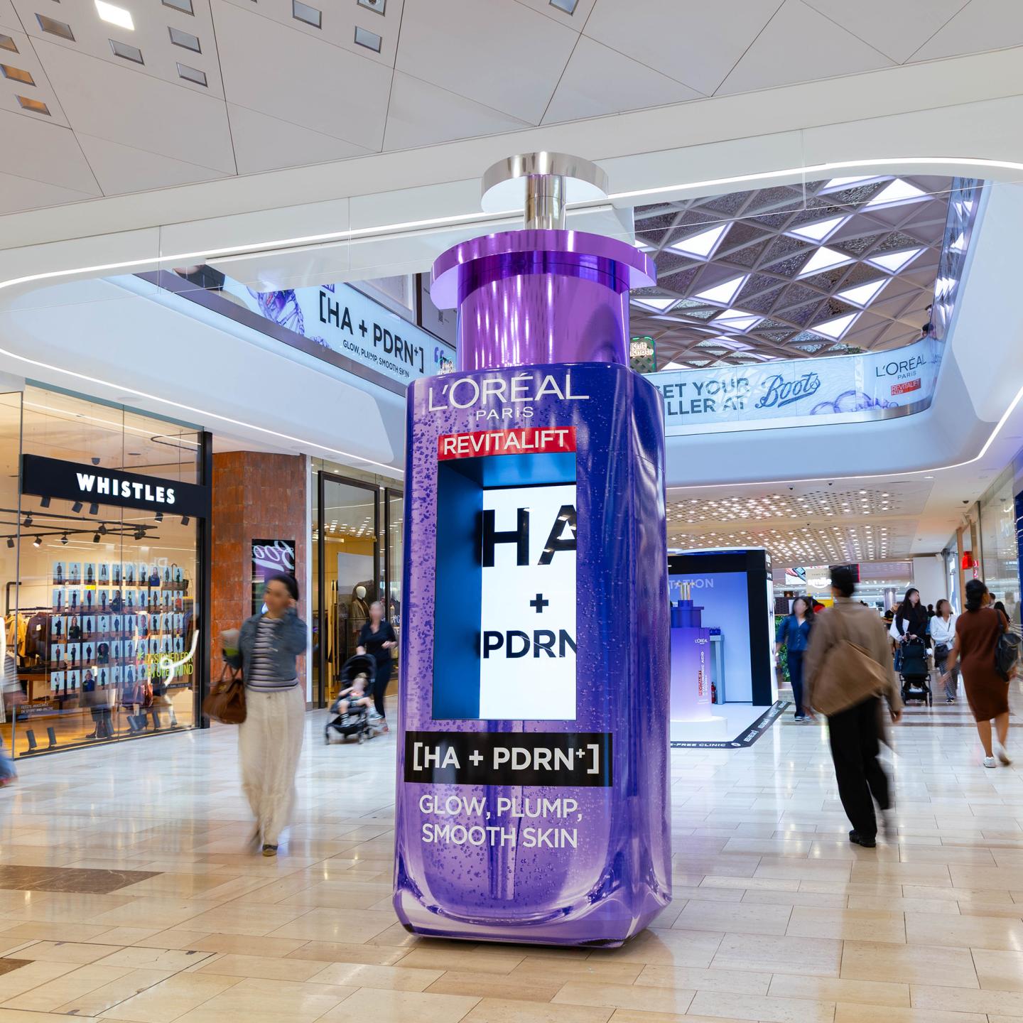 Tall purple 3D bottle of L’Oréal Revitalift [HA + PDRN] serum positioned in the central walkway of Westfield London shopping centre, with shoppers walking past fashion and beauty stores.