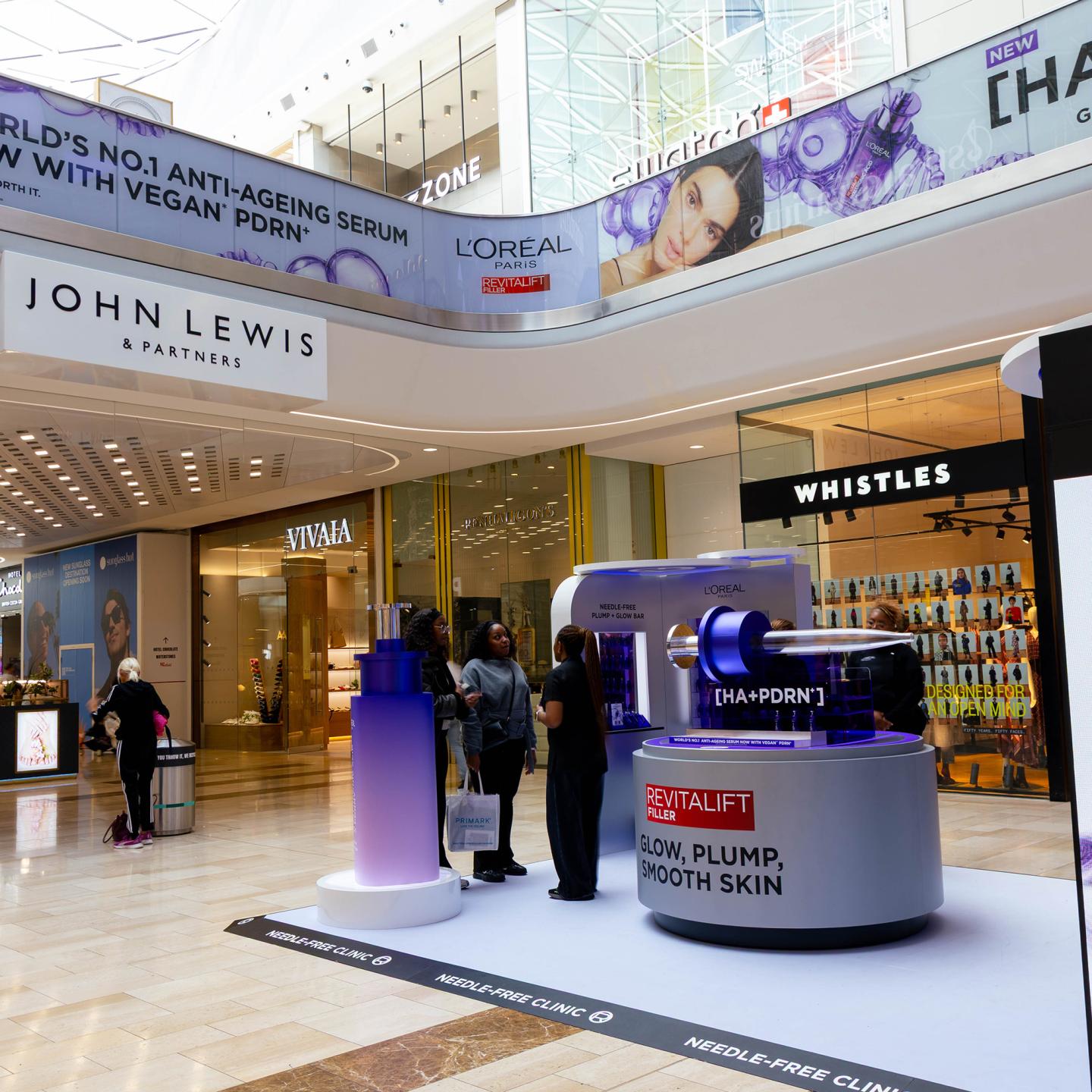 L’Oréal Revitalift experiential clinic setup inside Westfield London, with large anti-ageing banners above John Lewis and Whistles and shoppers engaging with the product display.