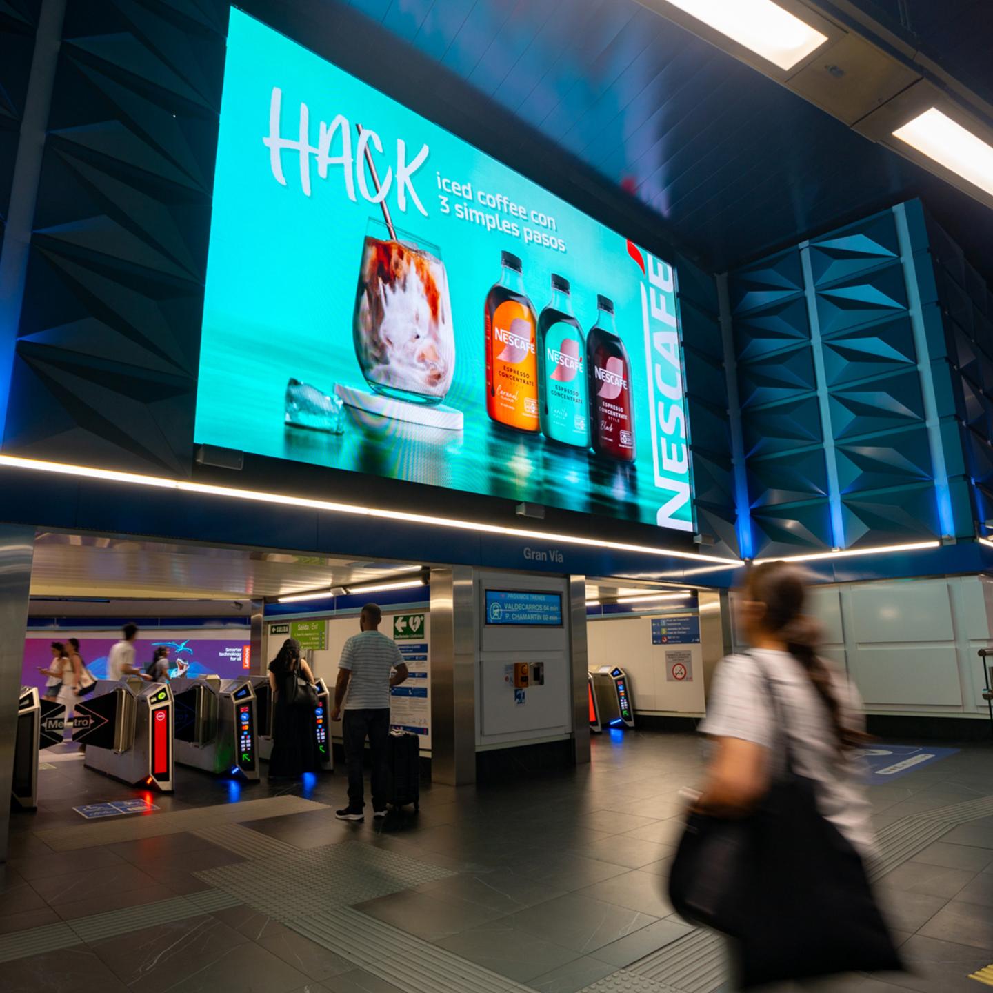 Large JCDecaux digital screen in Gran Vía metro station in Madrid displaying a NESCAFÉ Espresso Concentrate iced coffee advert above ticket barriers, with commuters walking through the concourse.