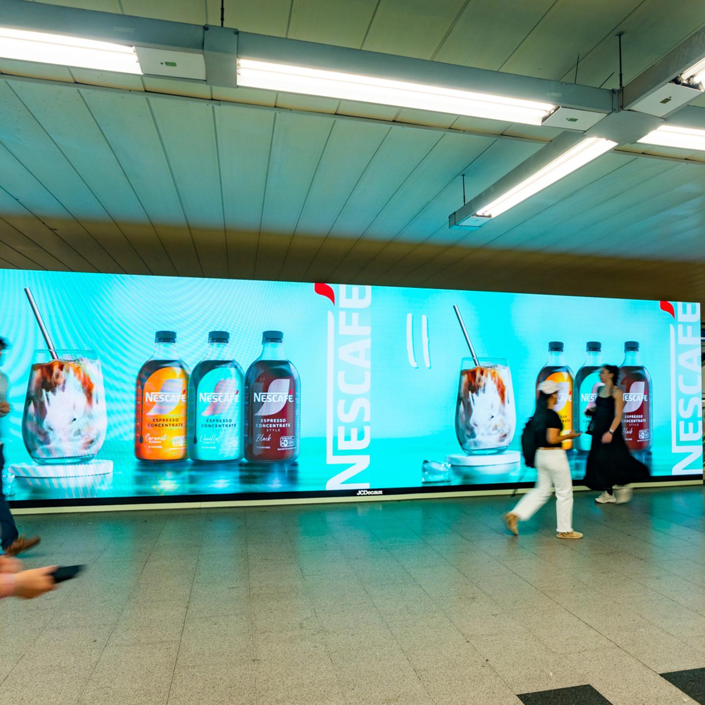 Wide JCDecaux digital wall in a Madrid metro corridor showing NESCAFÉ Espresso Concentrate bottles and iced coffee visuals on a turquoise background, with passengers passing in front of the screen.