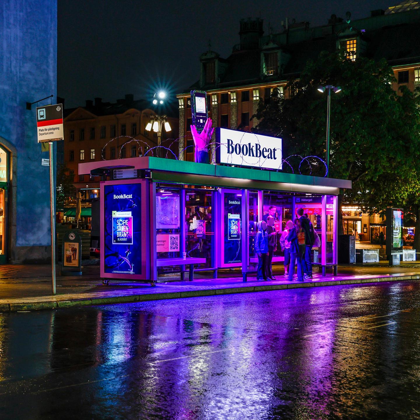 People waiting inside a JCDecaux Innovate Shelter wrapped in BookBeat branding, transforming the bus stop into an immersive audiobook experience.
