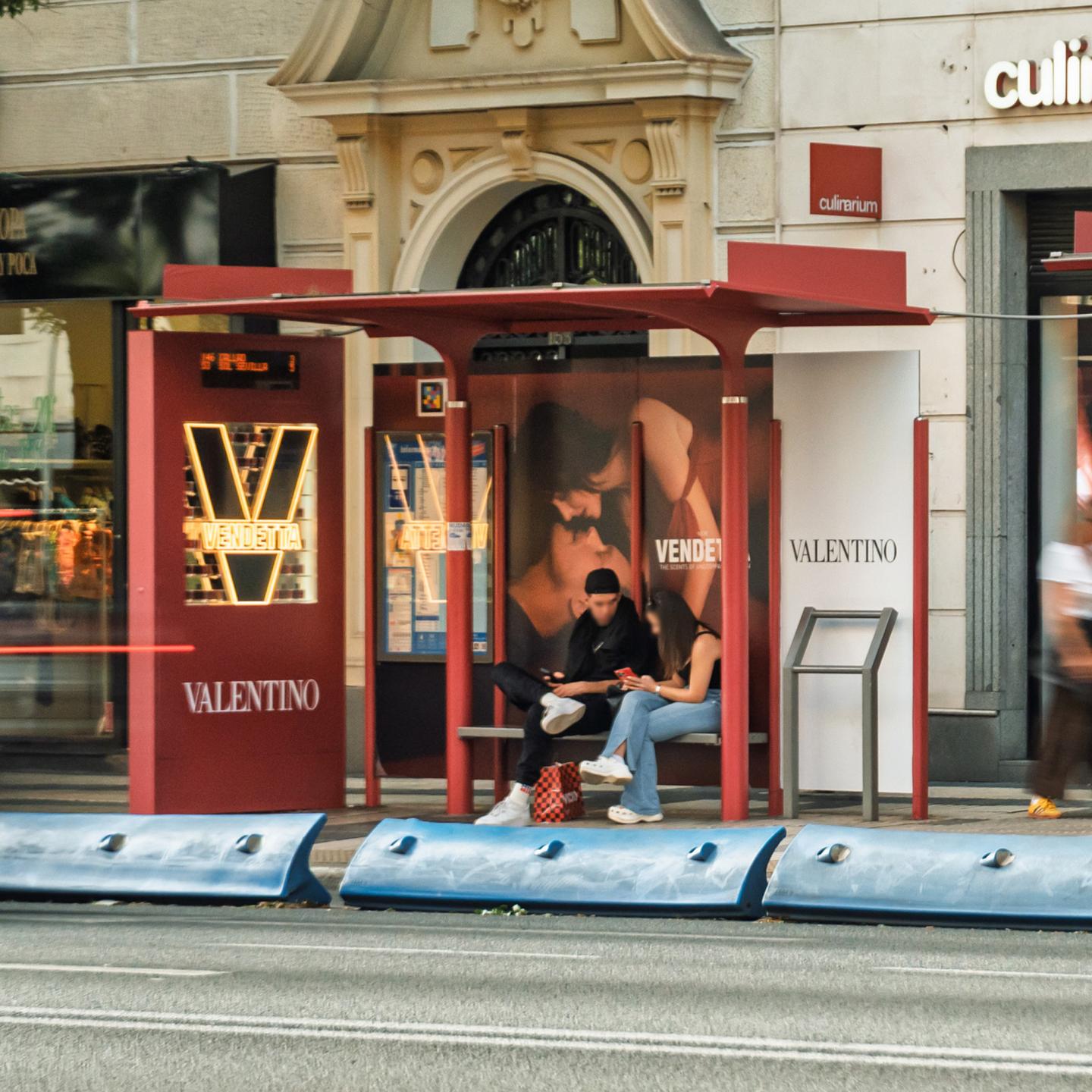 JCDecaux Spain bus shelter in Madrid fully branded in red for Valentino Vendetta, with two young commuters sitting beneath the illuminated perfume advert.
