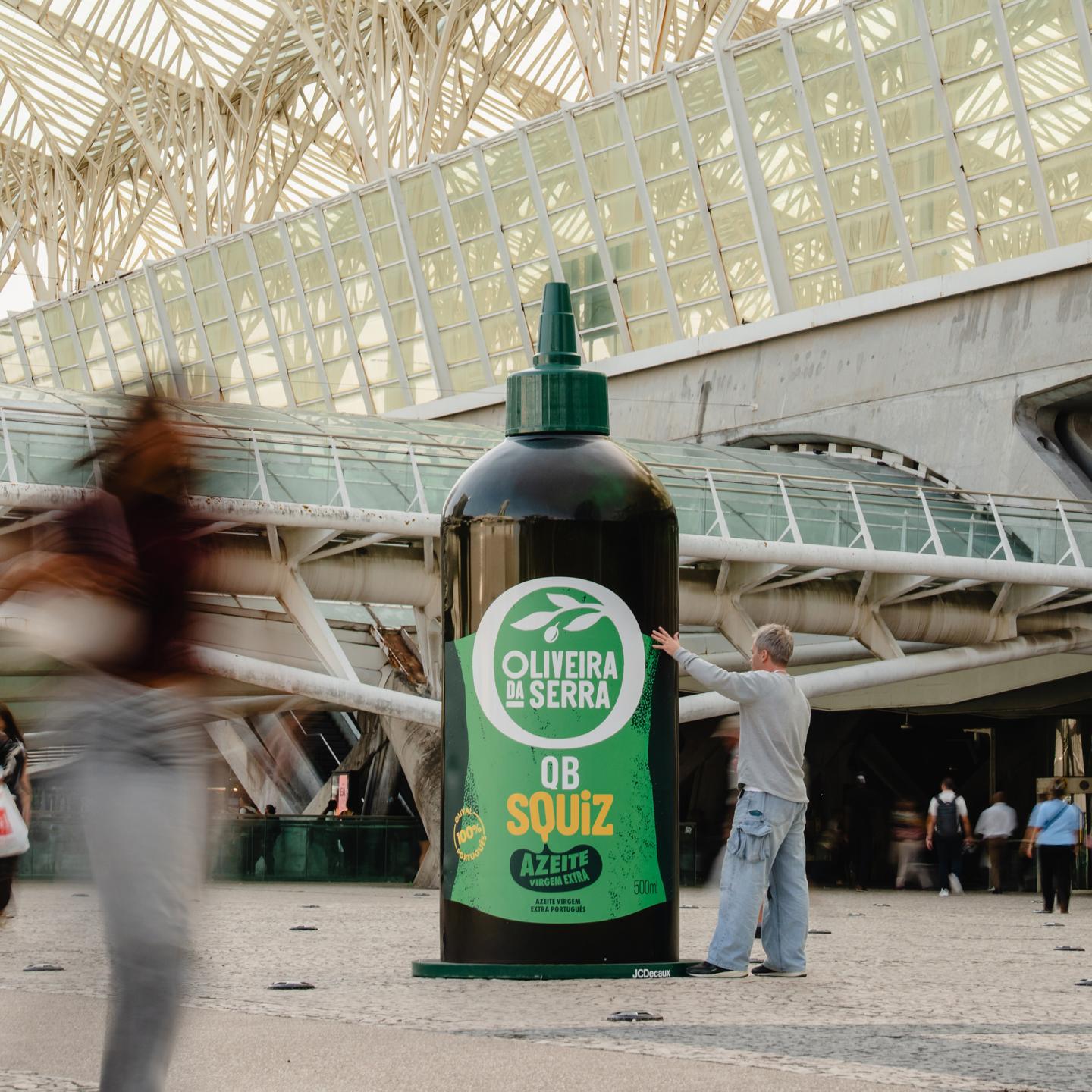 Close-up of an oversized Oliveira da Serra Squiz olive oil bottle in a busy Lisbon square, used as an eye-catching JCDecaux outdoor advertising installation.
